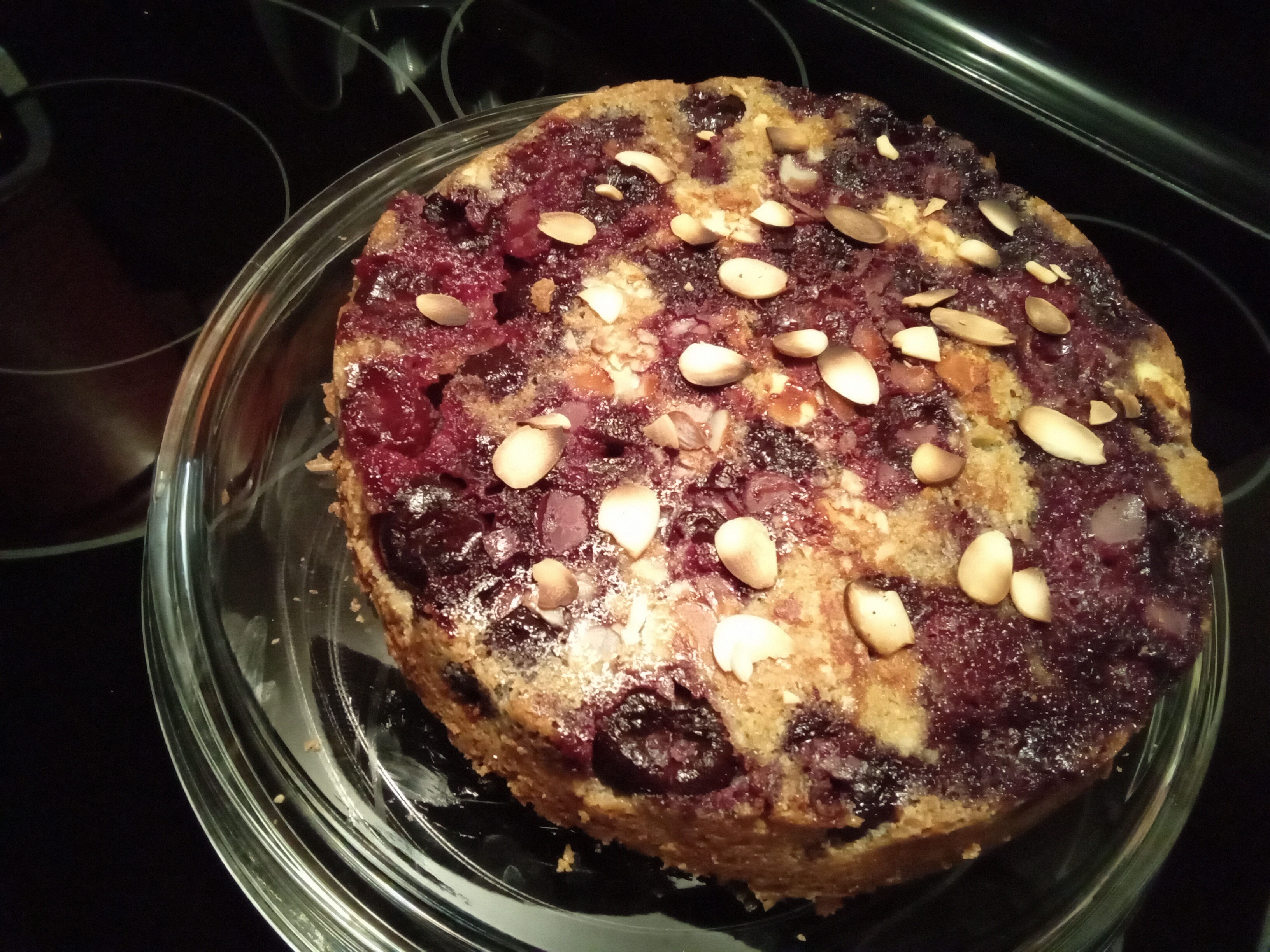 Vue en plongée d'un gâteau-pudding renversé, couverte de cerises cuites et d'amandes tranchées. La surface est striée entre rouge bourgogne des cerises et le brun doré du gâteau qui paraît plus qu'il ne devrait un peu.