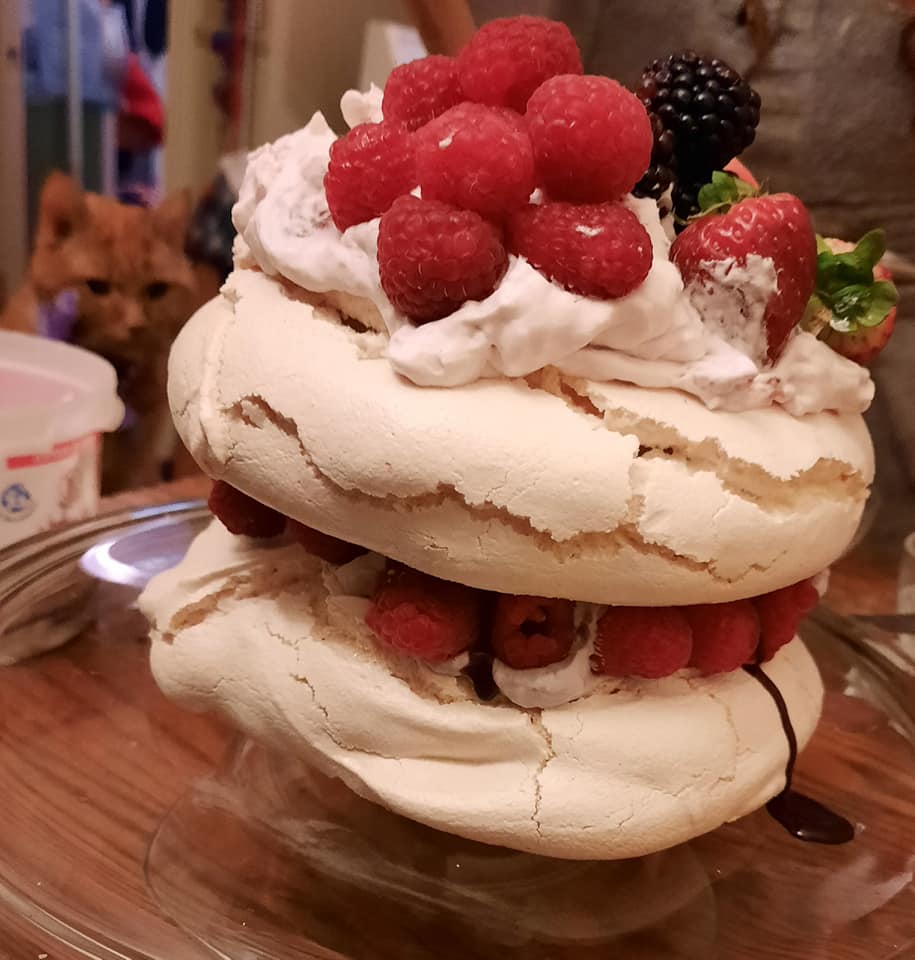 Une pavlova étagée couverte de fruits est en avant-plan, en arrière-plan on distingue les traits légèrement flous du visage de Clément fixant la pavlova. Ça doit être la crème fouettée qui le transfixe ainsi.