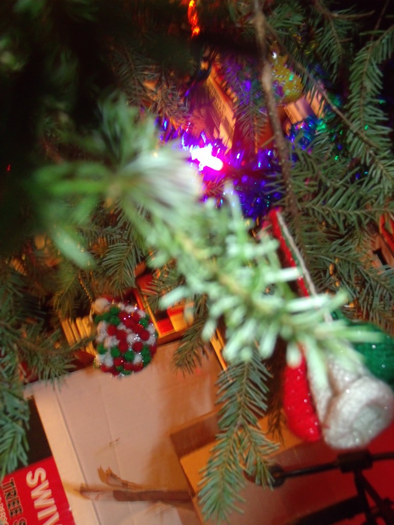 Vu de proche du dessus d'un de nos sapins passés, on y aperçoit vers la droite, les trois clochettes crochetées, accrochées ensemble à une branche, trio vert, rouge et blanc. Une boule de Noël de verre peinte est visible dans le coin supérieur droit, parmi les branches du sapin.