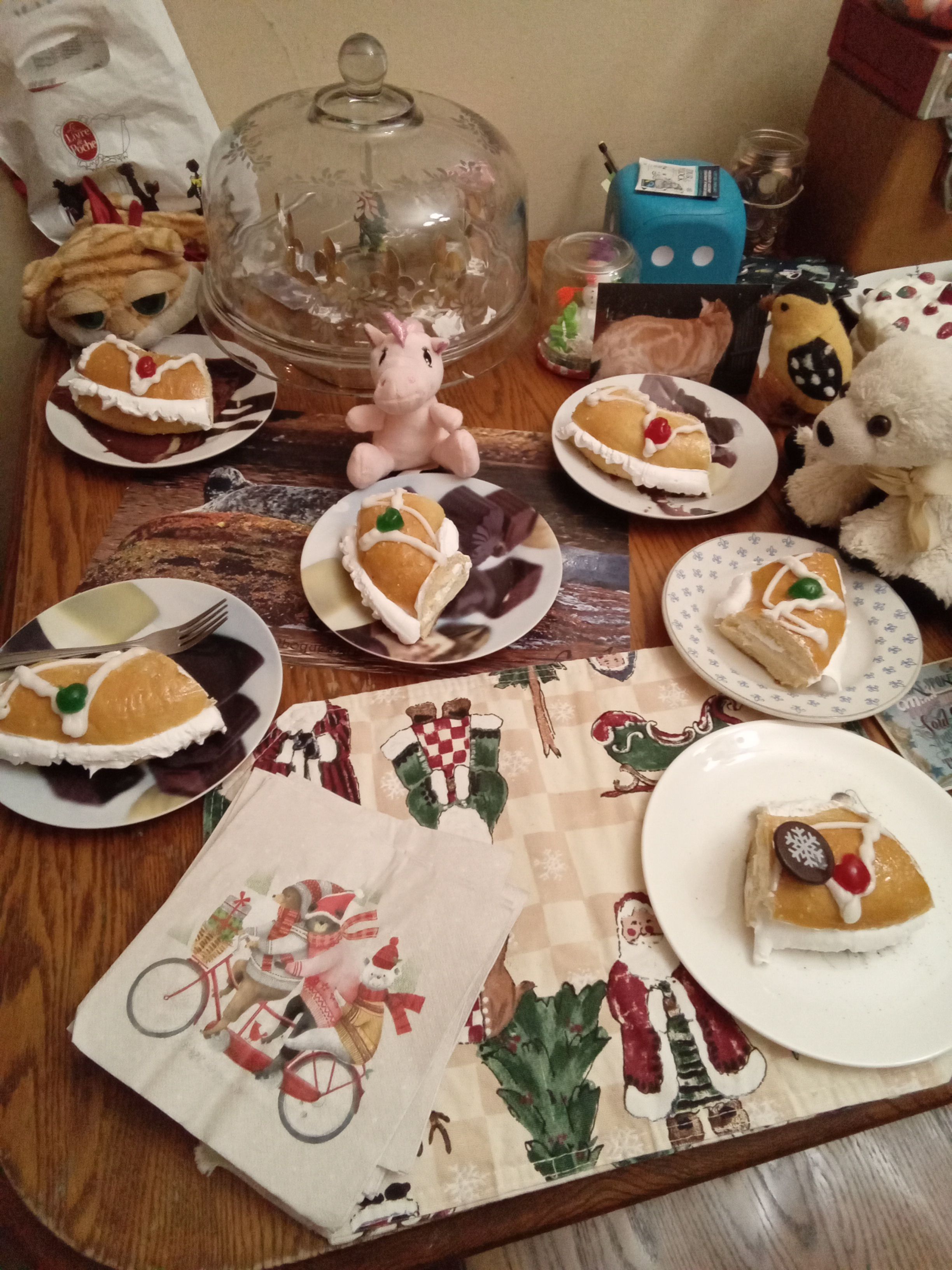 Six assiettes remplies de parts de couronne des rois briochées sont disposées sur la table, dont une devant Clément II la peluche chat tabby roux, Iconiki la peluche licorne rose, Paula la peluche ourse polaire de Clément, et une part devant la photo de Clément, gardé par une peluche feutrée d'un chardonnet jaune.