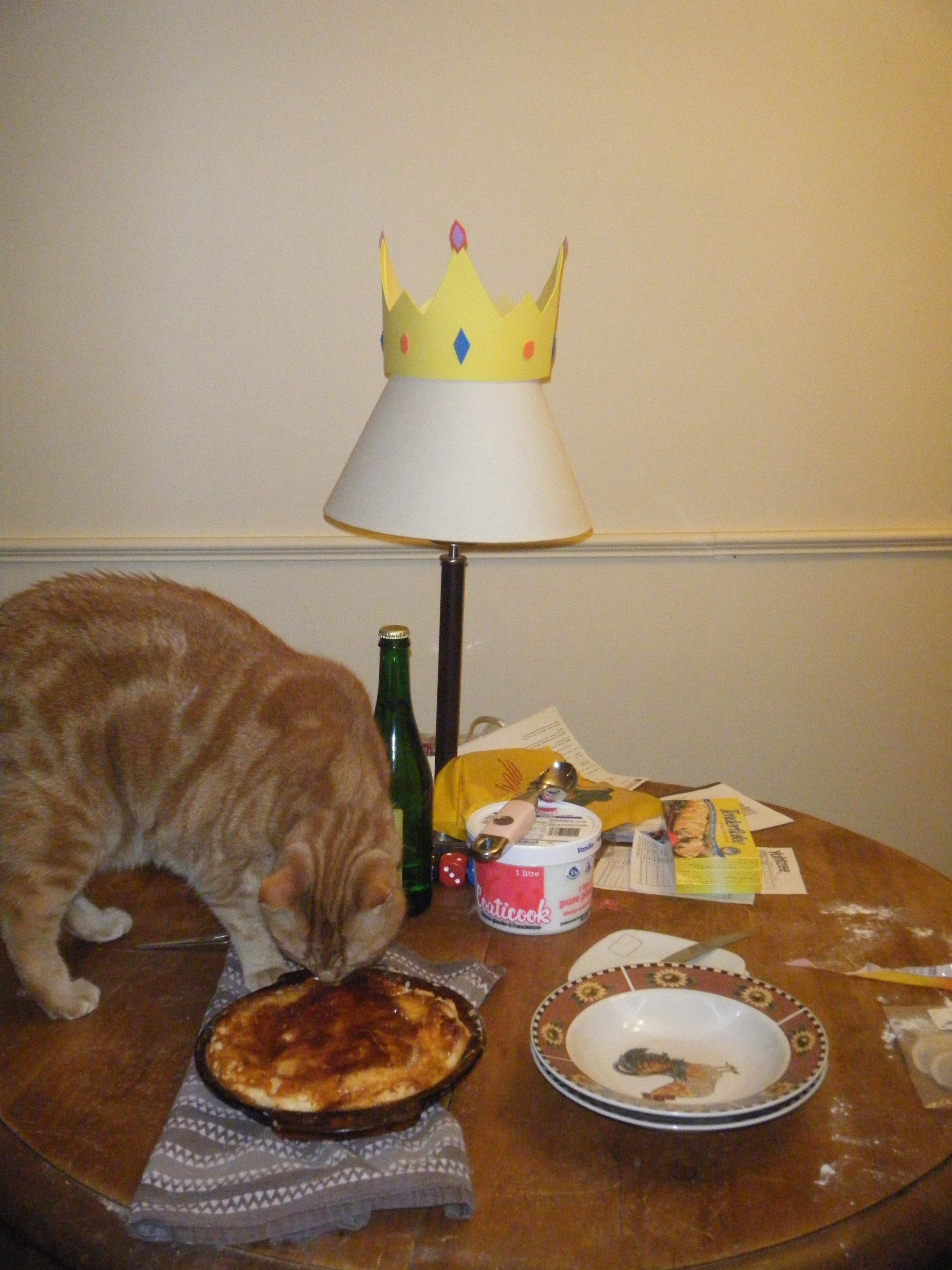 Clément le chat toujours debout sur la table est penché et semble humer d'un peu trop près la galette des rois à la frangipane légèrement trop cuite.