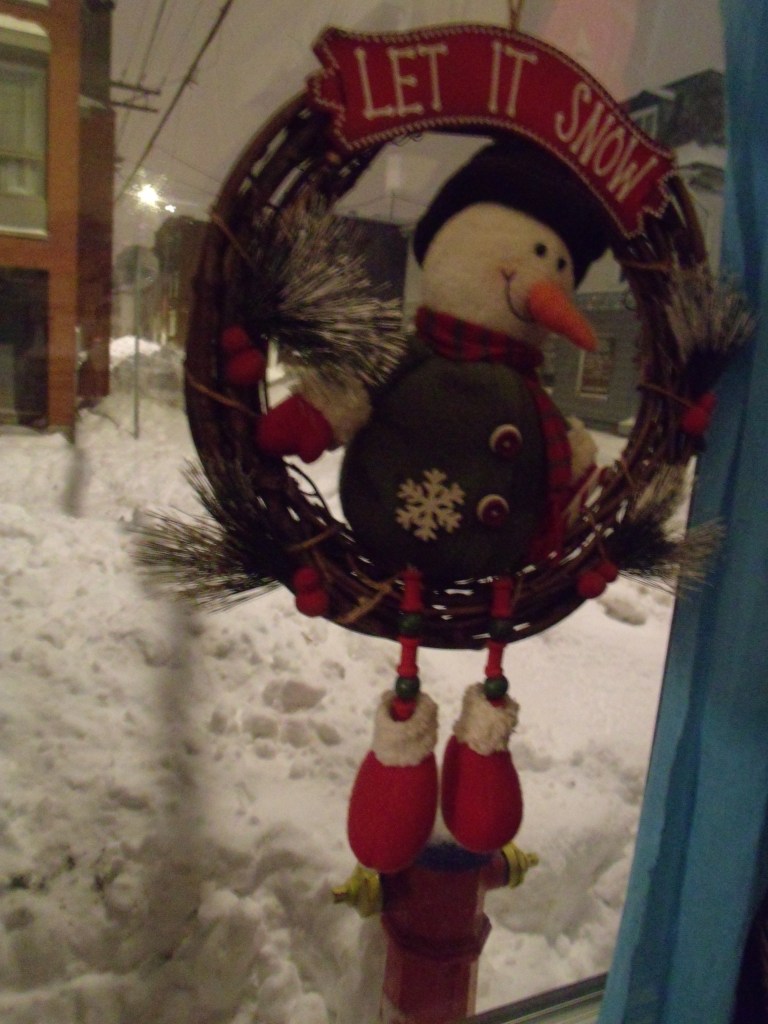 Une couronne de vignes avec un bonhomme de neige suspendue à la fenêtre, la rue derrière est pleine de neige.